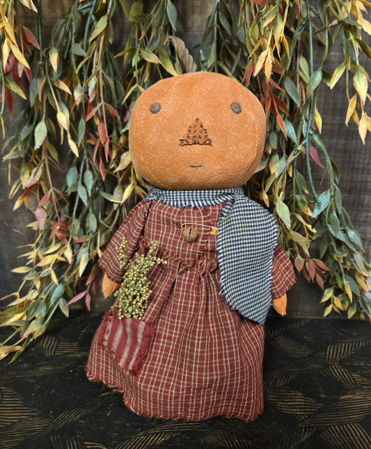 Textile art piece resembling a person with a brown head, plaid dress, and checkered scarf against a dark background with greenery.
