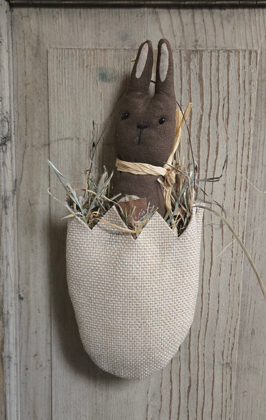 Textile rabbit figure in an egg-shaped basket on a wooden wall.