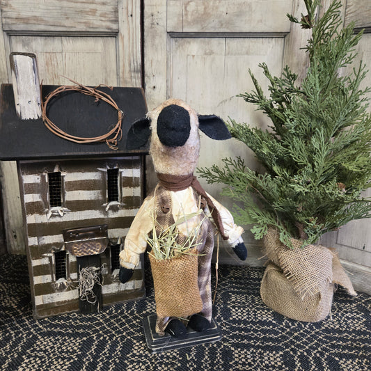A figurine of a sheep holding a bag of grass, wearing a brown shirt, with a decorative bow tie, placed on a wooden surface, with a green plant and a decorative item in the background.