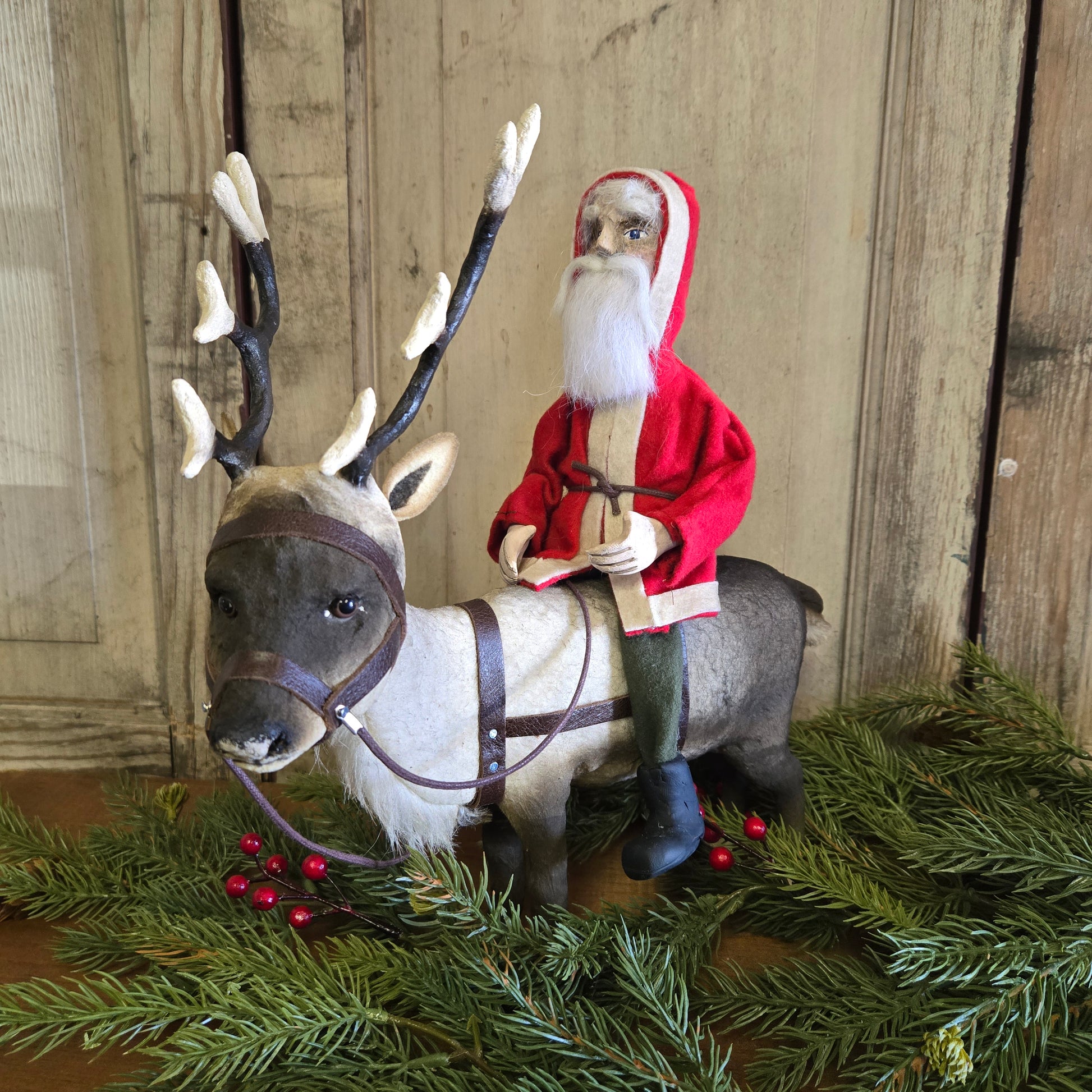 A handcrafted paper mache reindeer with a Santa Claus figure riding it, decorated with a red suit and white beard, positioned on a green pine branch with artificial snow and pine needles around.