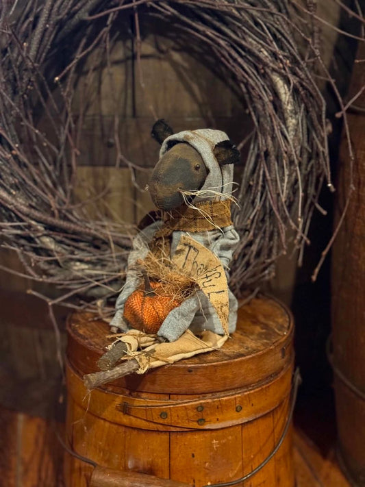 A rustic handmade rag doll with a gray face and hat, wearing a blue-gray dress and holding a small orange pumpkin, displayed on a wooden barrel with a twig wreath behind it.
