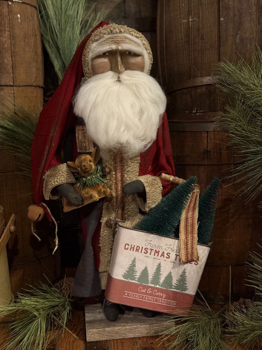 A rustic Santa Claus holiday figurine with a long white beard, red hat and coat, holding a small gingerbread man and a tin of Christmas trees, set against a wooden barrel backdrop with pine branches.