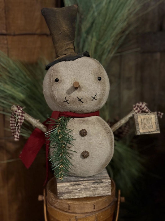A rustic handmade snowman decoration with a tall dark top hat, red scarf, green pine branch, and wooden base, shown against a dark holiday background with pine branches.