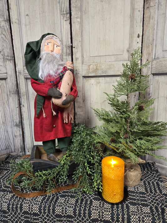 A figurine of Santa holding a snowman, with a red hat and coat, standing next to a decorative tree and a candle.