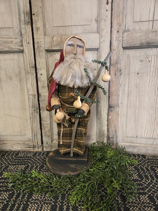 Primitive Santa holding a twig with gourds, standing on a base, with a plaid shirt and red hat, against a wooden backdrop.