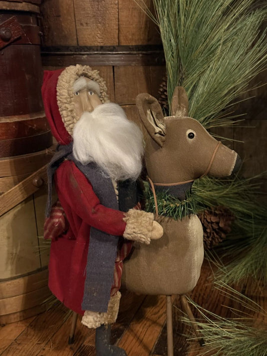 A rustic holiday decorative scene with a Santa Claus figure in a red coat and white beard riding a tan reindeer, set against wooden barrels and green pine branches.