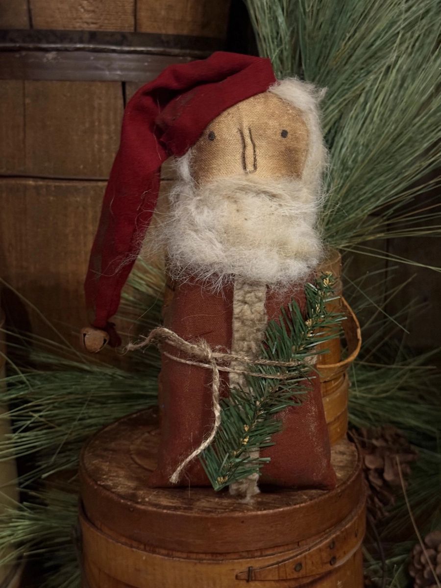 A rustic Christmas decorative figure resembling Santa Claus, made from a wooden face with white faux fur for a beard, wearing a red hat and brown body, displayed on a wooden barrel with pine branches in the background.