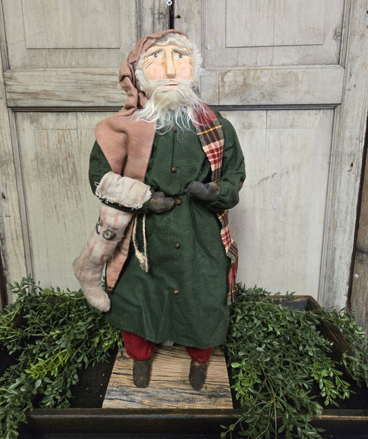 A clay face Belsnickel figure with a stocking and tan cap, dressed in green, placed on a wooden surface with holly and greenery in the background.