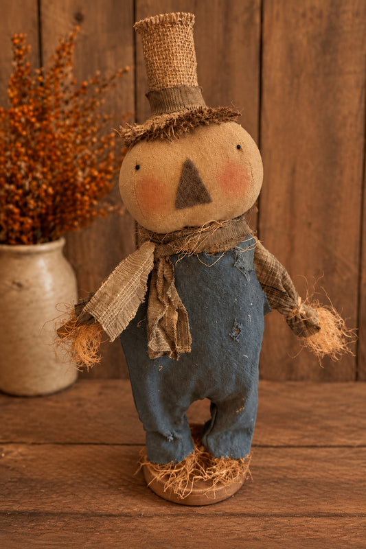 A rustic handmade fabric scarecrow doll with a burlap hat, blue denim-like pants, tan scarf, and straw-colored hands and feet, standing on a wooden surface against a wood plank background.
