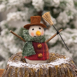 Decorative snowman figurine with a hat, scarf, and broomstick on a wooden stump with a blurred snowy background.