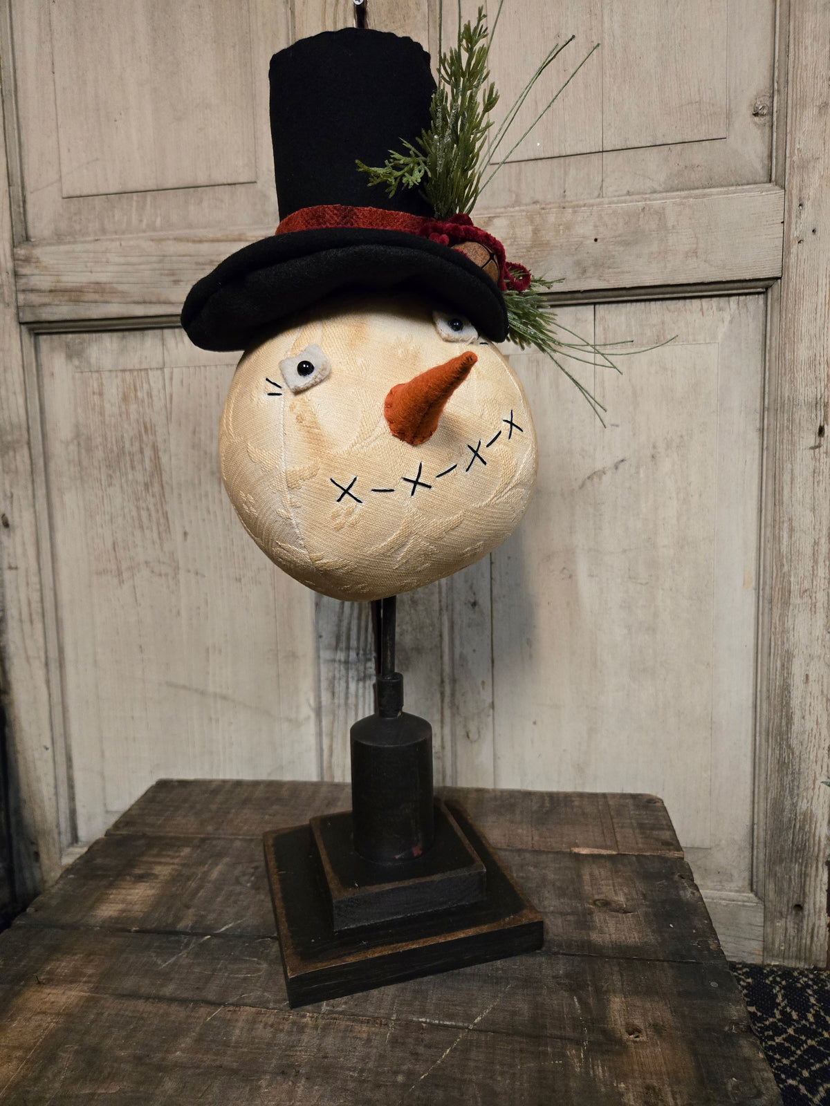 A decorative snowman head sculpture on a black stand, wearing a black top hat with a red band and green pine sprigs, set against a rustic white paneled door and wooden table.