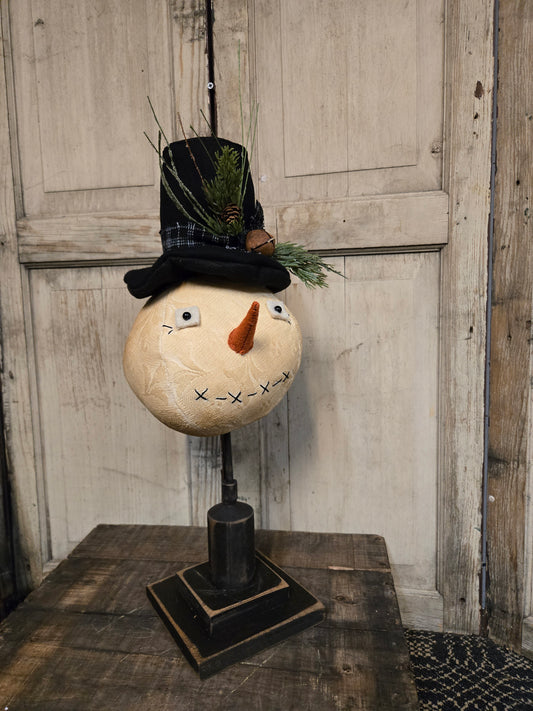 A rustic decorative snowman head sculpture on a black metal stand, wearing a black top hat with green pine branches and a small pinecone, set against a weathered wood door background.