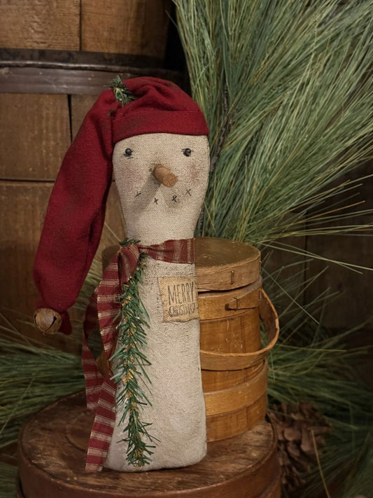 A rustic Christmas snowman decoration made from a beige fabric bag, wearing a red Santa hat and a red-and-white striped scarf with green pine garland, displayed on a wooden surface with a wooden bucket and evergreen branches in the background.