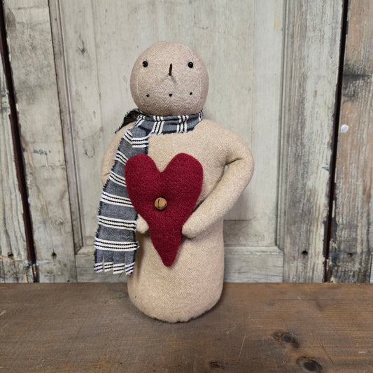 A beige handcrafted primitive snowman with a red heart-shaped detail and a grey scarf, standing against a wooden backdrop.