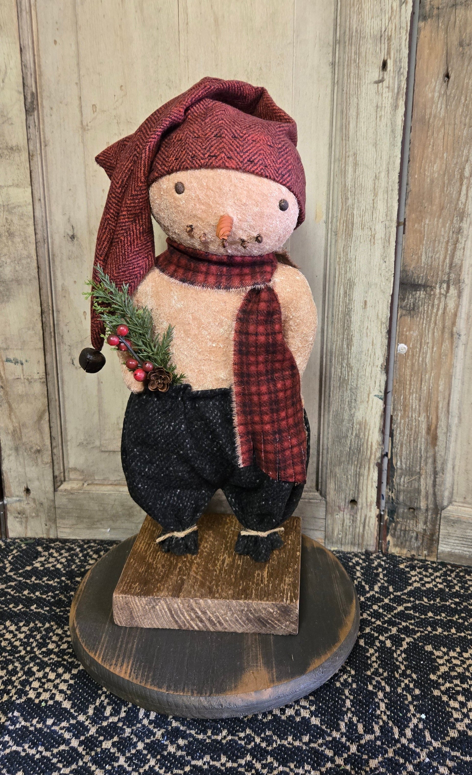 Snowman doll dressed in red and black with a sprig of pine displayed on a black wooden riser