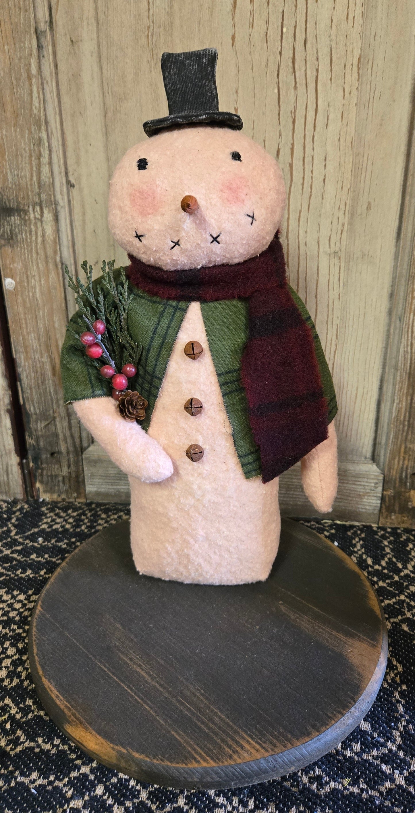 A 12-inch handcrafted snowman with top hat made of cotton batting and flannel attire, adorned with pinecones and berries, standing on a wooden base.
