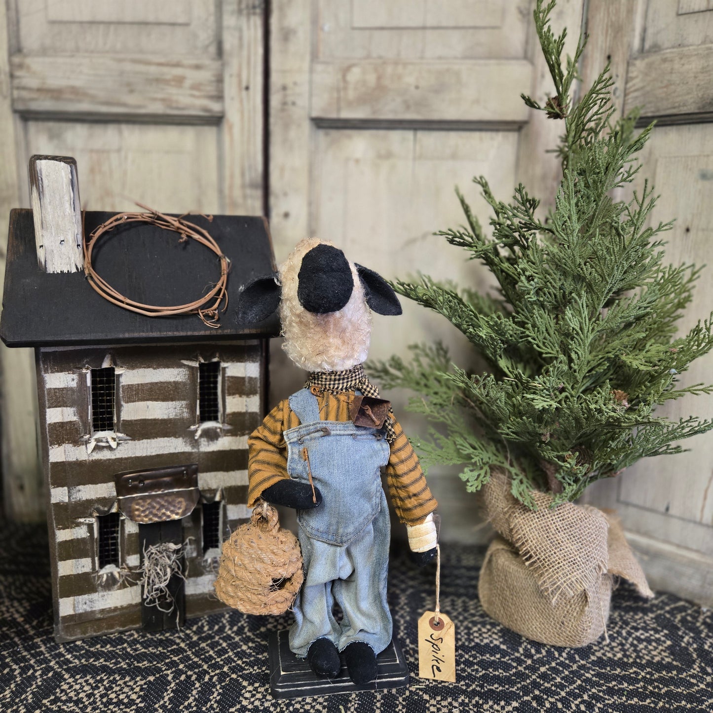 A handcrafted figurine of a sheep wearing overalls and holding a beeskep, displayed on a rustic-themed setup with a wooden cabinet, a pine cone, and a burlap sack.