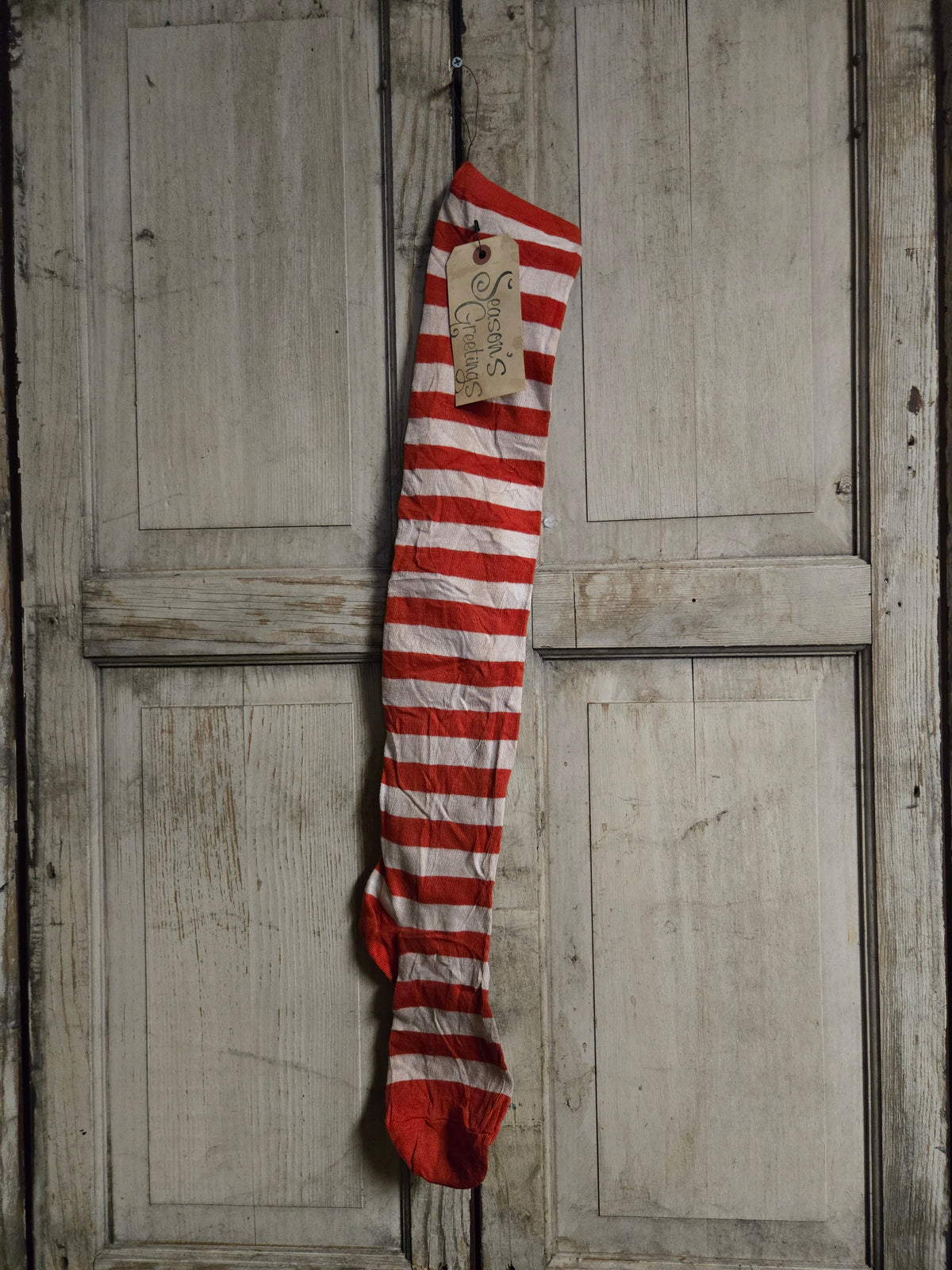 A single red and white striped sock hangs vertically against a distressed, light gray wooden door. A small paper tag is attached near the top.