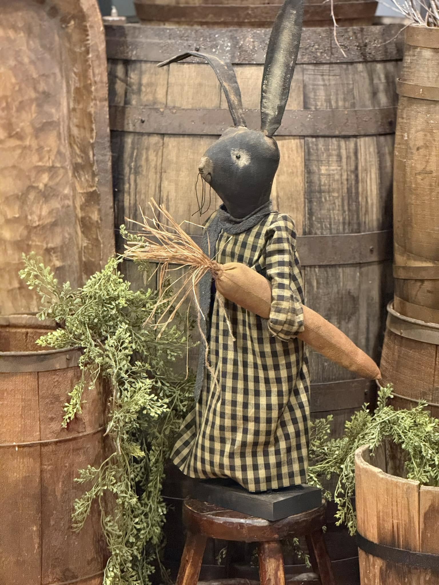 A decorative figurine of a primitive style rabbit dressed in a black and tan checked dress, holding a large carrot, positioned against a rustic backdrop.