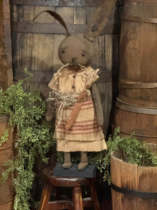 A plush toy resembling a brown rabbit wearing a grungy muslin dress and a burgundy ticking apron, standing against a rustic backdrop.