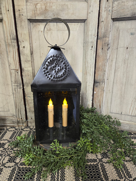 A black tin large ship's lantern with a handle on top and two candles inside, placed on a table with a patterned tablecloth. The lantern is set against a backdrop of a wooden door and wall.
