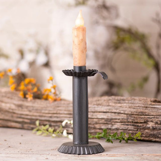 Tin candle holder to accommodate a real or battery operated taper candle with flowers in background.