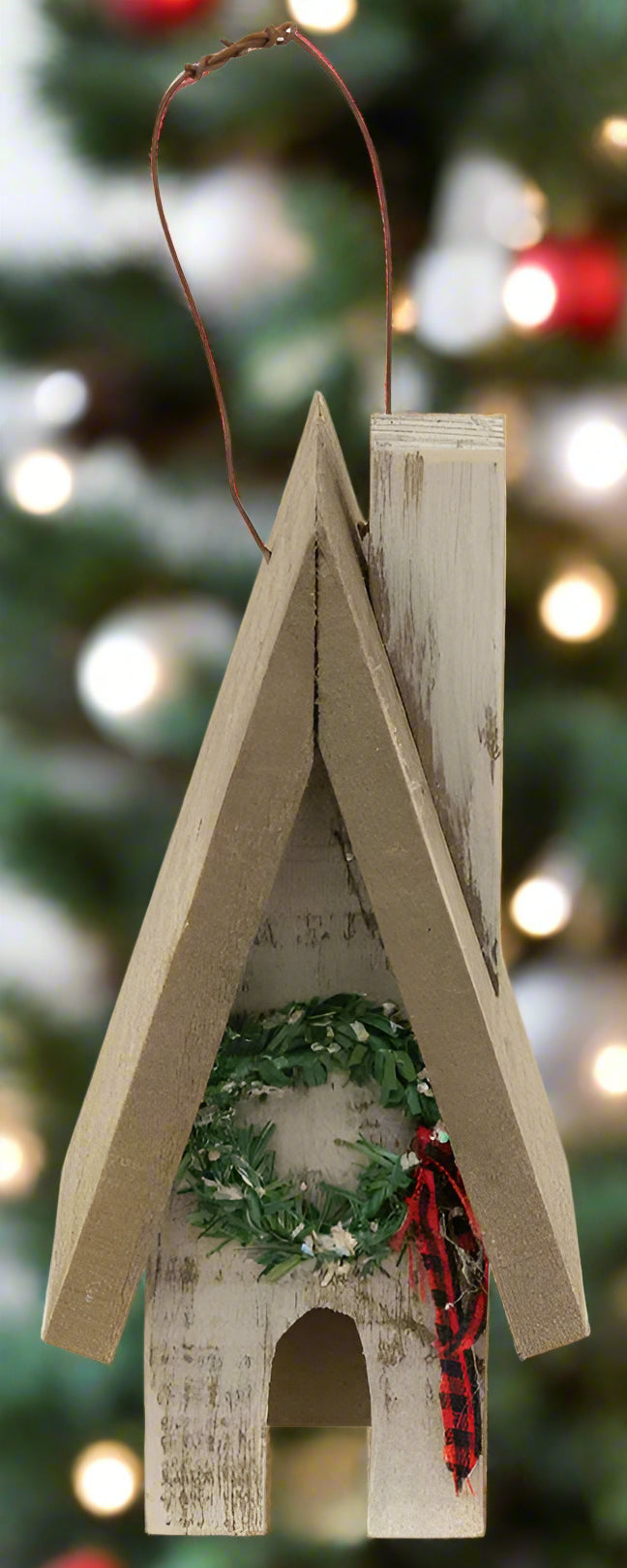 A wooden ornament in the shape of a town house with a snowy wreath accent hanging above the door, and a wire hanger attached at the top.