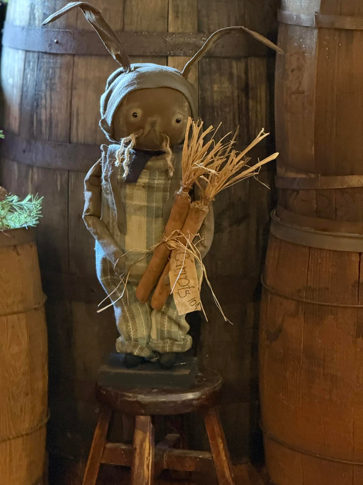 A handmade Primitive Wallace Bunny doll with a flannel bib, plaid shirt, and hat, holding a bundle of wheat, displayed in a rustic setting.