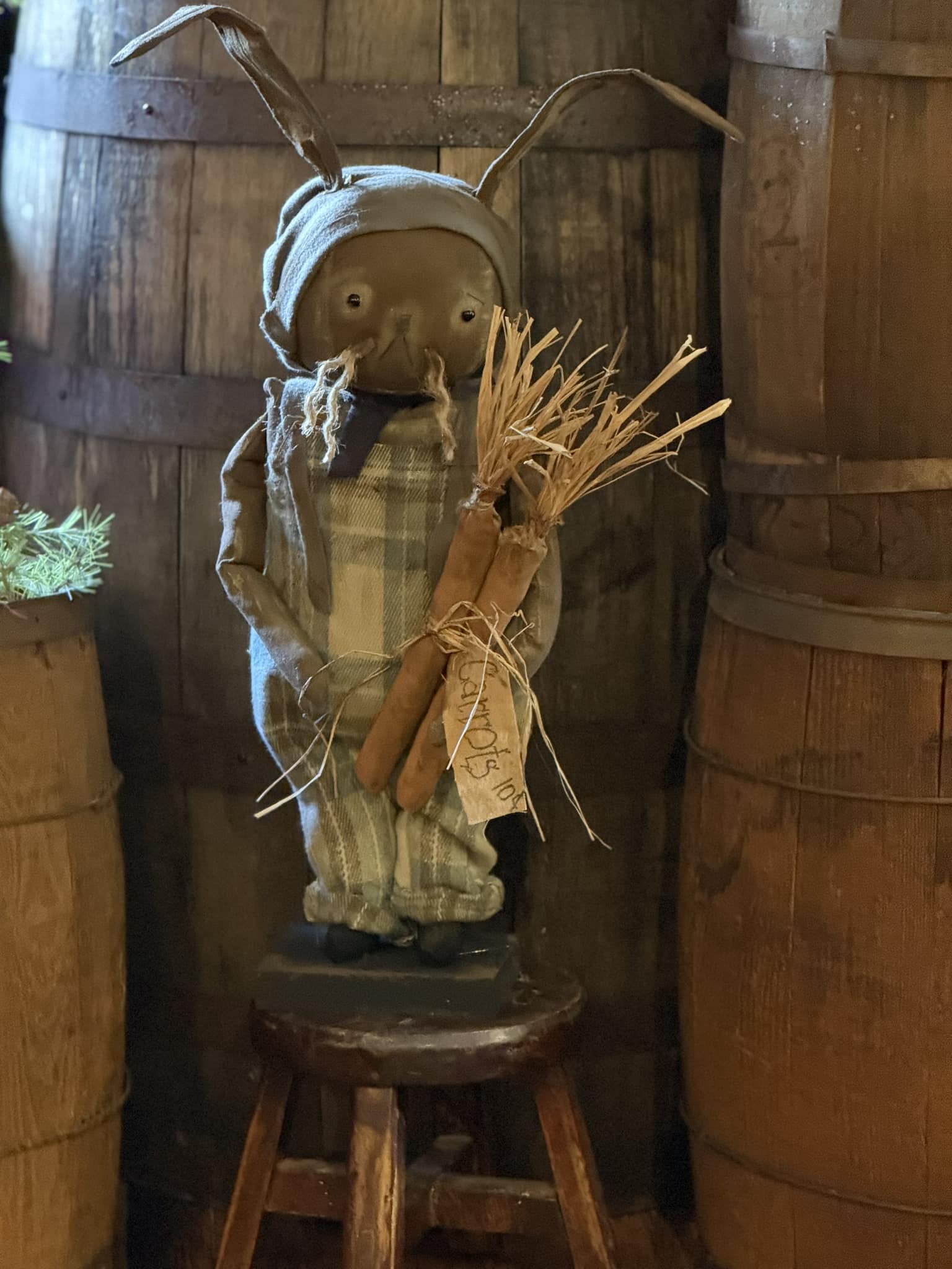A handmade Primitive Wallace Bunny doll with a flannel bib, plaid shirt, and hat, holding a bundle of wheat, displayed in a rustic setting.