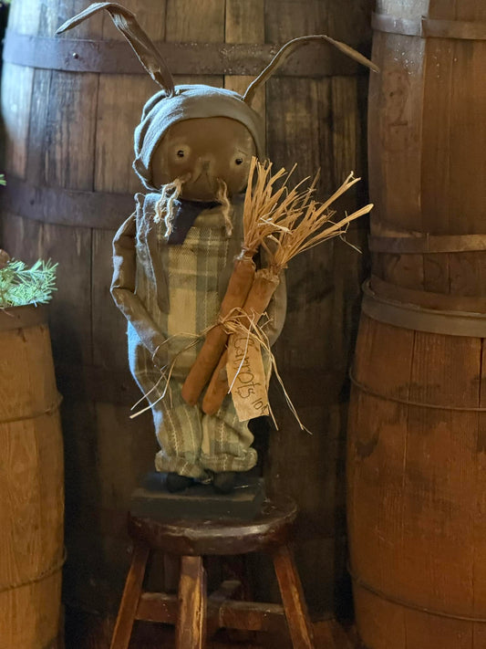 A handmade Primitive Wallace Bunny doll with a flannel bib, plaid shirt, and hat, holding a bundle of wheat, displayed in a rustic setting.