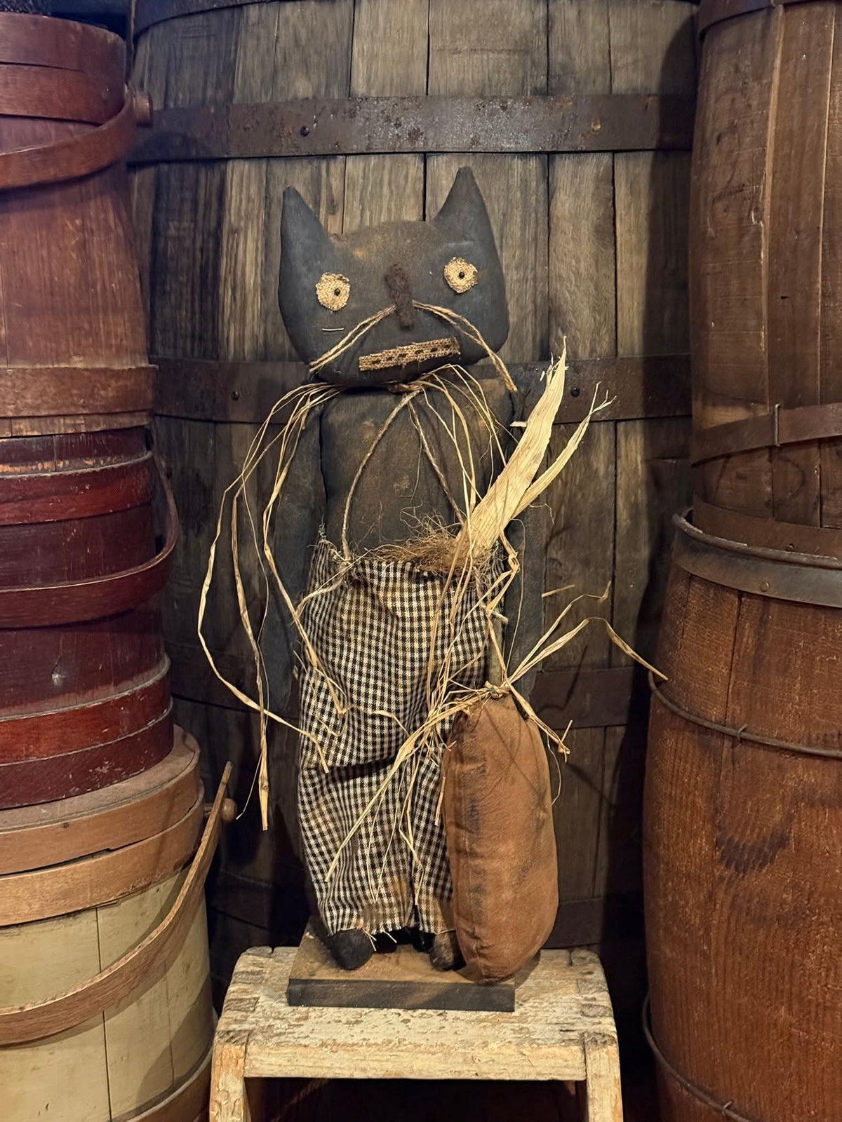 Vintage wooden cat figure with checkered shirt and corn cob, surrounded by wooden barrels.