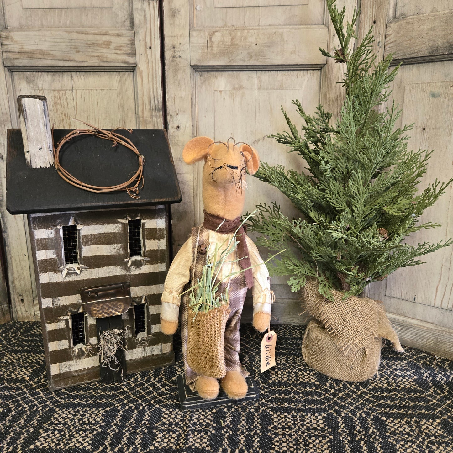 A handcrafted figurine of a mouse holding a bag of grass, wearing a plaid outfit, positioned next to a miniature house and a decorative greenery bundle.