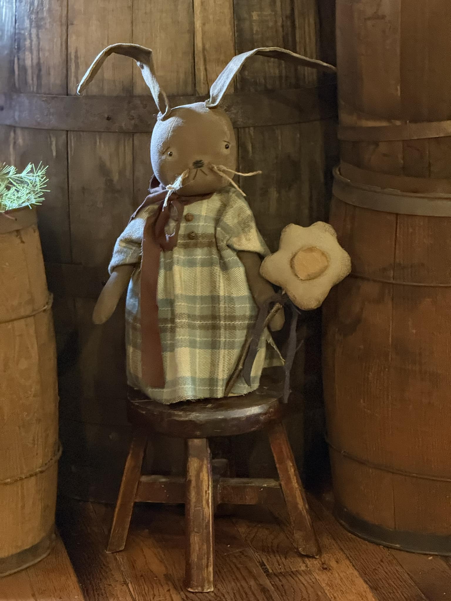 A plush toy resembling a bunny dressed in a checkered dress and holding a flower, seated on a wooden stool against a backdrop of wooden barrels.