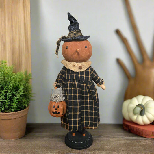 Halloween-themed doll with a witch outfit, pumpkin basket, and black hat on a wooden surface.