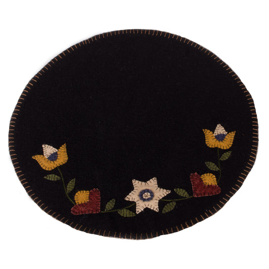 A round, black wool felt mat with a floral flowering vine design and a visible yellow, blue, and red stitched border.
