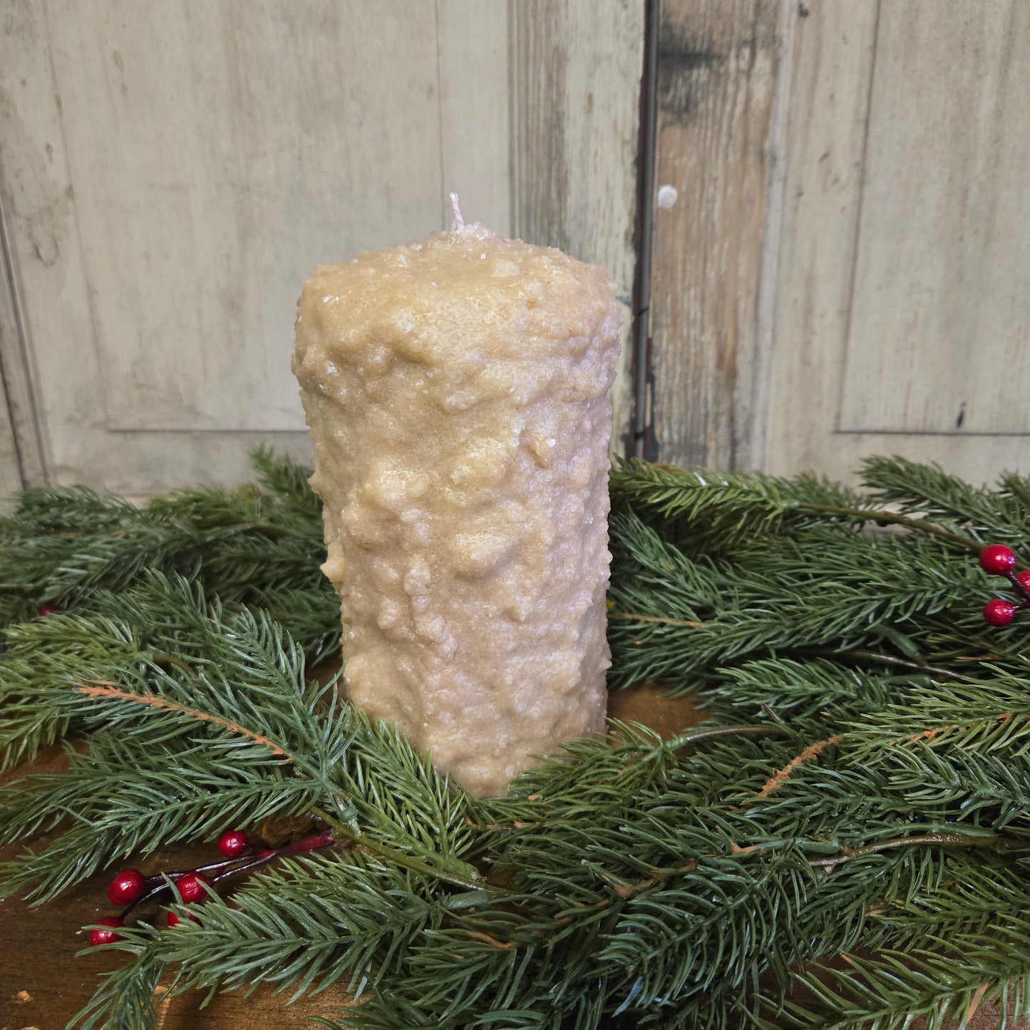 A large pillar candle with a cranberry center, covered in brown sugar and cinnamon, placed on a decorative wreath with green pine branches and red berries.