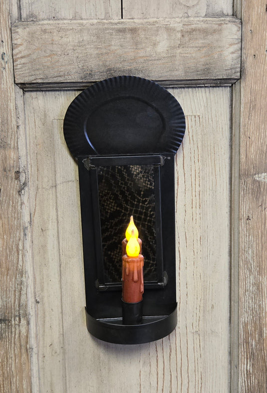 A black tin crimped mirrored sconce mounted on a wooden wall, with a candle-like light source visible through the mirror.