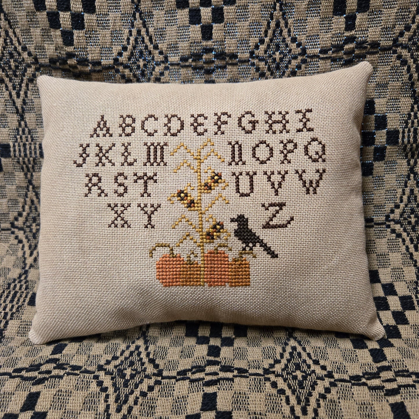 A square beige cross-stitch pillow featuring the alphabet and a small bird design, placed on a patterned blue and beige textile background.