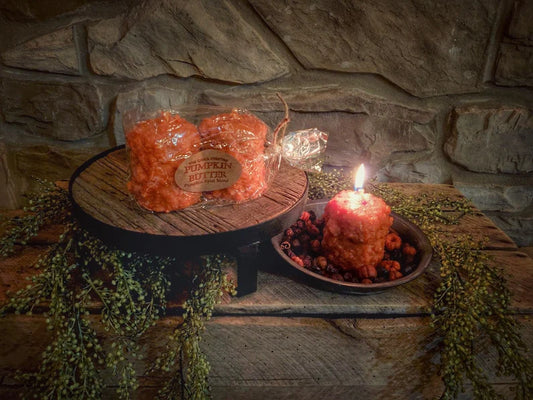 Two pumpkin-colored candles on a wooden platter with a label in the center, surrounded by decorative stones and greenery, with a lit candle on the side.