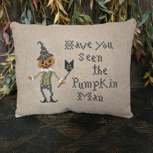Decorative pillow with cross-stitched design of a pumpkin man and text, surrounded by autumn leaves.