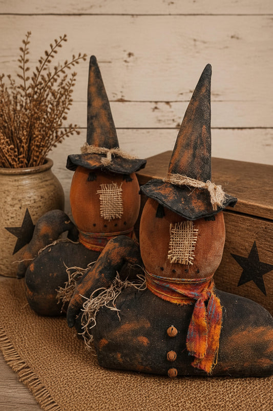 A rustic Halloween decorative figure shaped like a seated figure wearing tall pointed witch hats, a burlap face patch, and an orange plaid scarf, styled with dried grass in a ceramic pot and a wooden box on a burlap runner. The text 'Primitive Kreations Wholesale' appears in the upper right.
