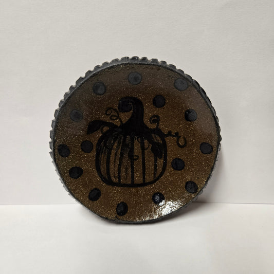 A round, textured dark brown and black decorative plate featuring a black pumpkin silhouette in the center and black polka dots around the border, set against a plain light background.