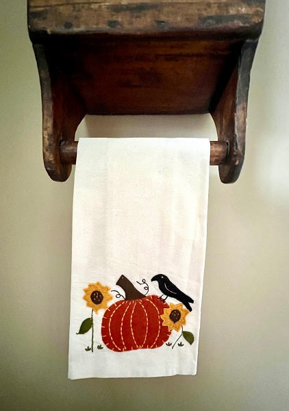 A white hand towel with a pumpkin and a crow design, hanging on a wooden towel bar.