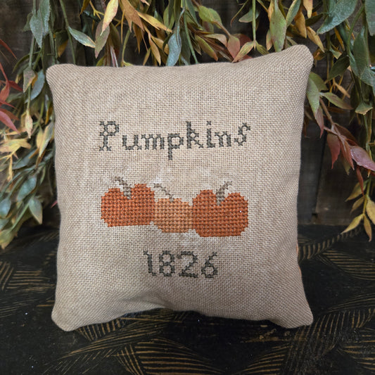 Decorative pillow with cross-stitched pumpkin design and text on a leafy background