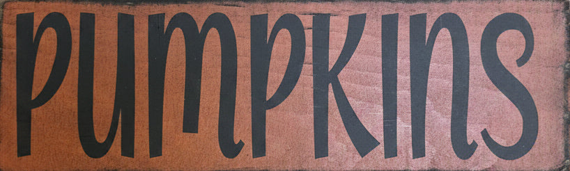 A rustic vertical wooden sign with the word 'pumpkins' in large black script on a warm brown wood background, shown against a light weathered wood plank backdrop.