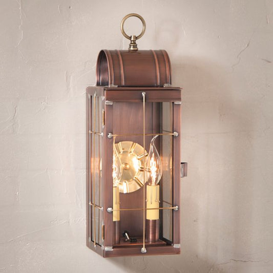 An antique copper Queen Arch lantern with a clear glass enclosure and brass-tone hardware, featuring two light bulbs inside, mounted on a wall.