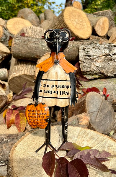 Decorative figure with a pumpkin and text, standing on a log with woodpile and leaves in the background