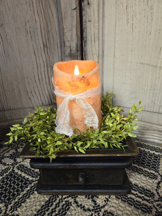 A realistic moving flame 5-inch pillar candle in burnt ivory color, placed on a decorative tray with greenery around it.
