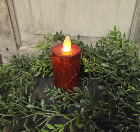 A realistic moving flame votive candle with a burgundy wax column surrounded by green foliage.