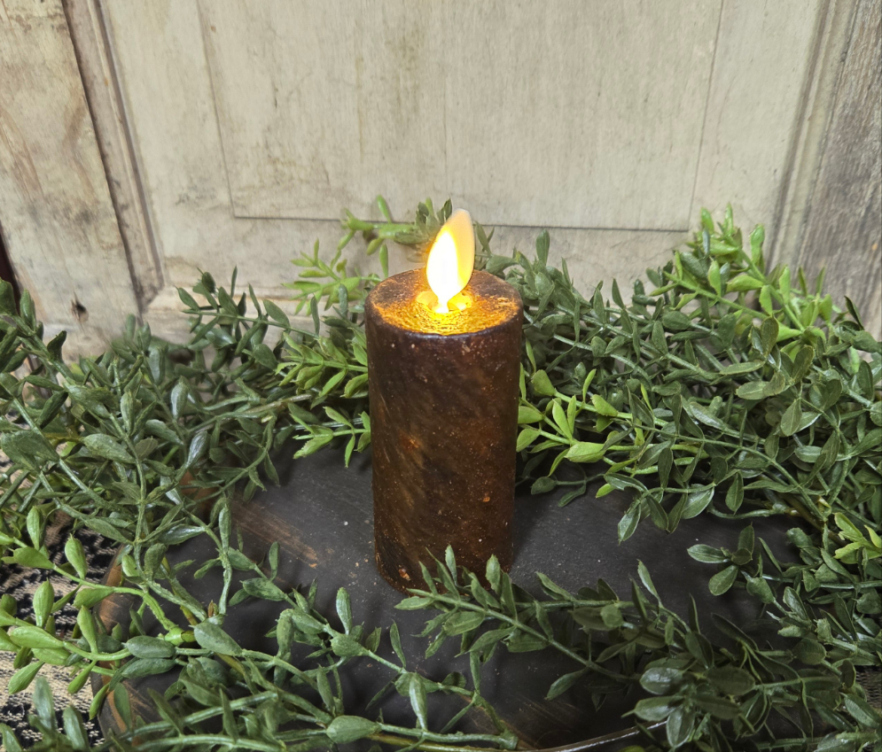 A realistic moving flame votive candle with a dark brown color, placed on a dark surface surrounded by greenery. The candle appears to be made of wax with a realistic appearance.
